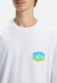 Weißes Baumwoll-T-Shirt mit einem bunten Grafikdesign im linken Brustbereich in Blau- und Grüntönen. Rundhalsausschnitt, kurze Ärmel.