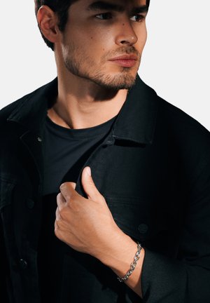 Black denim jacket over a black shirt, with a silver chain bracelet on the wrist. Strong facial features and defined jawline.