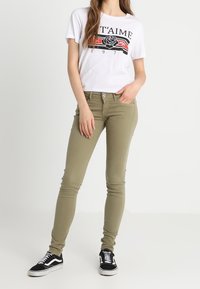 Jeans ajustados verdes combinados con una camiseta gráfica blanca que presenta un diseño de rosa negra, llevados con zapatillas negras y blancas.