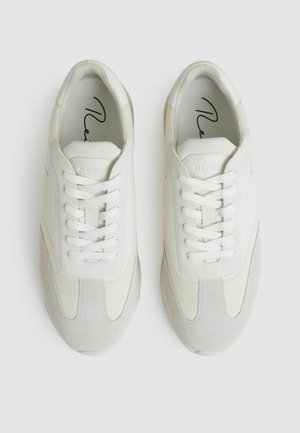 Zapatillas blancas que combinan materiales lisos y texturizados, acentos grises, puntera redonda y cordones. Diseño minimalista.