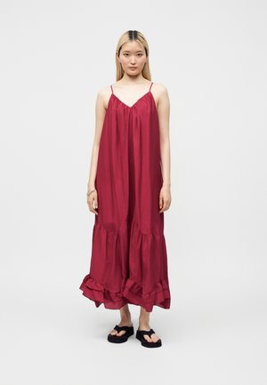 Junge Frau mit glatten blonden Haaren, die ein lockeres, ärmelloses rotes Maxikleid und schwarze Sandalen trägt, steht vor einem schlichten weißen Hintergrund.