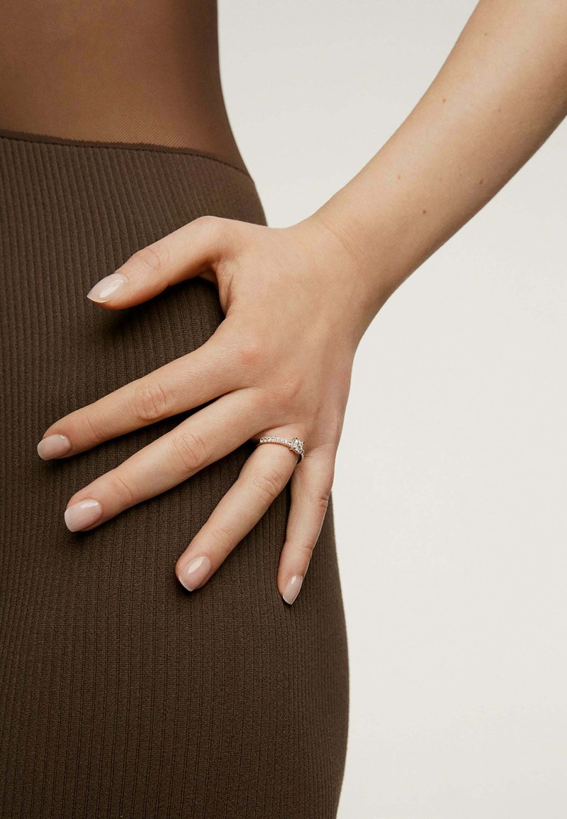 Anillo de plata con múltiples pequeñas piedras preciosas, llevado en una mano apoyada sobre una tela marrón con textura acanalada. Un esmalte de uñas neutro complementa el look general.