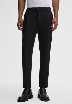 Black, tailored trousers with a smooth texture, pleated front, elastic waistband, and a button closure; worn with black shoes.