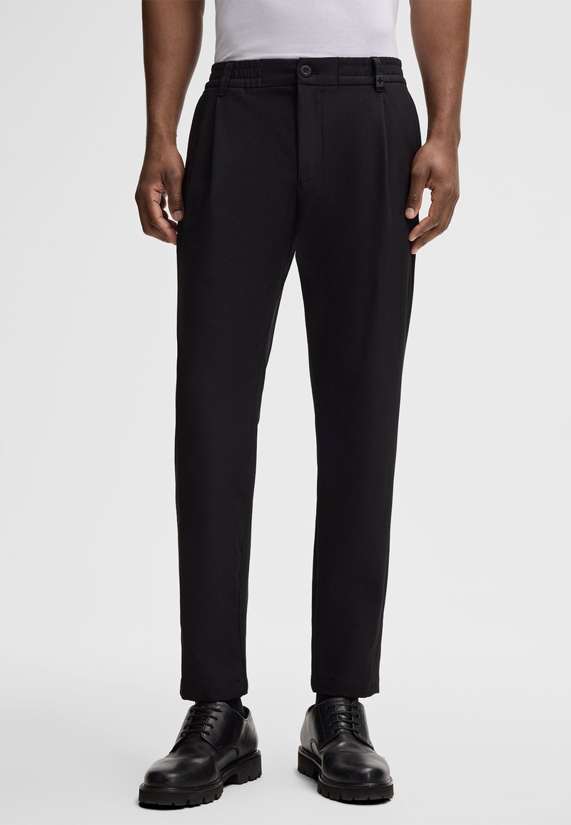 Schwarze, maßgeschneiderte Hose mit glatter Textur, gefaltetem Vorderteil, elastischem Bund und Knopfverschluss; getragen mit schwarzen Schuhen.