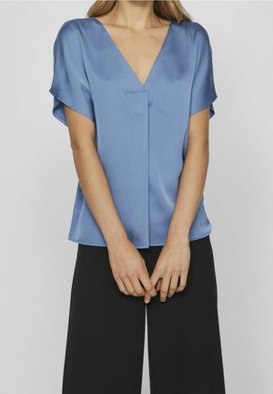 Femme portant un chemisier en satin bleu clair à manches courtes et col en V, avec un pantalon noir ample, debout, les mains jointes devant elle.