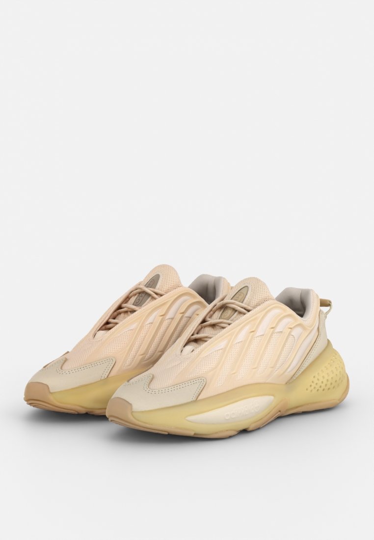 Zapatillas deportivas de un tono beige claro con parte superior de malla, detalles texturizados y suela acolchada con una forma curva y un diseño minimalista.