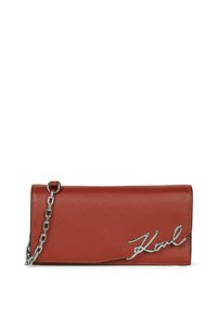 Cartera de cuero rojo con una textura suave, que cuenta con una correa de cadena plateada y un acento metálico con la firma. Forma compacta y rectangular.