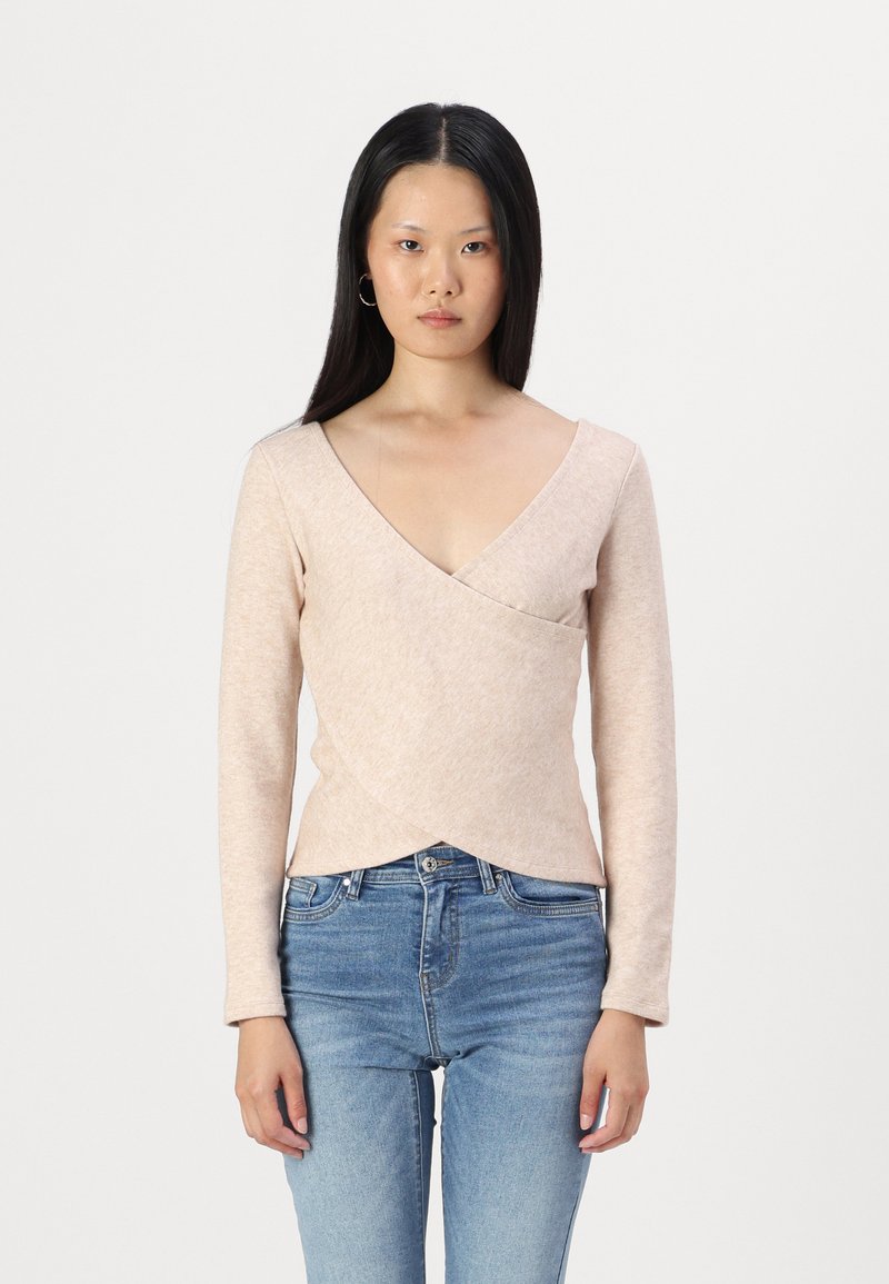 Hellrosa, langärmliges Wickeltop aus weichem Stoff mit V-Ausschnitt und tailliertem Design, kombiniert mit blauen Jeans.