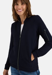 Giacca a zip blu marina realizzata in tessuto a coste con colletto alto, tasche laterali e tasca con zip sulla manica. Presenta un motivo testurizzato.