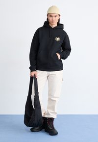 Sort hættejakke med et logo, cremede stribede bukser og sorte snørebodssko. Modellen holder en sort skuldertaske. Blå gulv baggrund.