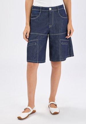 Donna che indossa pantaloncini di jeans blu scuro fino al ginocchio con tasche frontali e scarpe bianche Mary Jane, in piedi contro uno sfondo bianco.