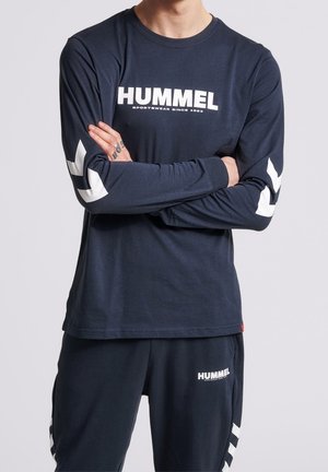 Granatowa koszula z długim rękawem z białym logo i brandingiem "HUMMEL", z białymi akcentami w kształcie zygzaka na rękawach. Wykonana z miękkiego materiału bawełnianego.