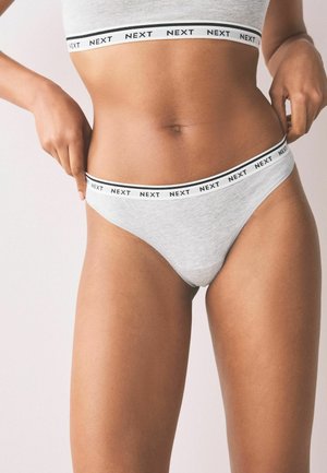 Detail osoby v šedém spodním prádle a podprsence s elastickými pásky s nápisem "NEXT", která si upravuje pas.