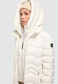 Jachetă albă tip puffer cu glugă mare, design matlasat, închidere cu fermoar în față și detaliu cu logo negru pe mânecă. Textură moale și netedă.