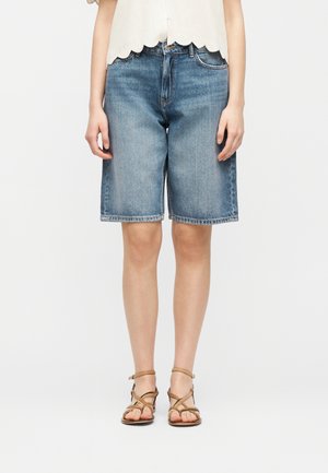 Femme portant un short en jean bleu mi-long, un haut blanc à bords festonnés et des sandales plates à brides marron clair, debout devant un fond blanc.
