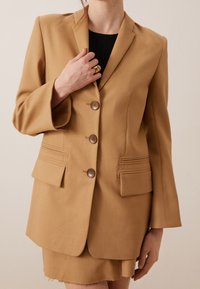 Blazer sur mesure couleur noisette avec trois boutons, revers crantés et deux poches avant. Tissu lisse avec des accents de couture verticale.
