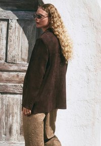 Blazer en daim marron au style oversize, associé à un pantalon beige texturé. Le modèle a de longs cheveux ondulés et porte des lunettes de soleil rondes.