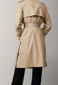 Beżowy płaszcz trenchowy z paskiem w talii, tylnym klapą i dopasowanym krojem. Tkanina wydaje się gładka i lekka.
