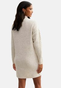 Frau mit dunklem Haar, die ein cremefarbenes, geripptes Strickkleid trägt, steht mit leicht gedrehtem Rücken und schaut zur Seite.