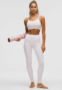Gaiši rozā sporta krūšturis un legingi, gluda tekstūra, pieguļošs dizains, kreisajā rokā turēts sarullēts rozā un baltas krāsas jogas paklājiņš.