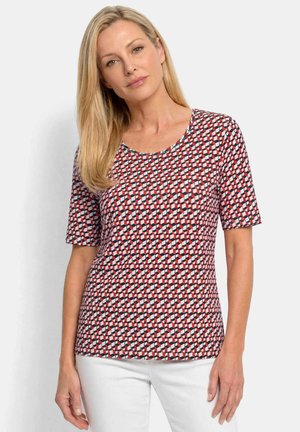 Blondine draagt een top met korte mouwen met rood-wit-zwart geometrisch patroon en een witte broek, staand tegen een effen achtergrond.