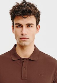 Brązowa koszulka polo z textured tkaniny, z kołnierzykiem, zapięciem na dwa guziki oraz subtelnym logo na lewej piersi.