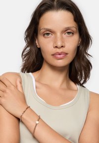 Roze gouden armband om de pols met kralen en een ronde bedel. Het model draagt een lichtgroene tanktop met een witte binnenafwerking.