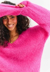 Puszysty, oversizeowy różowy sweter z dekoltem w kształcie litery V, długimi rękawami i miękką teksturą. Noszony przez osobę o kręconych włosach.