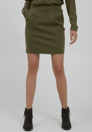 Femme portant une jupe vert olive et un haut assorti avec des bottines noires, debout avec une main dans la poche devant un fond uni.