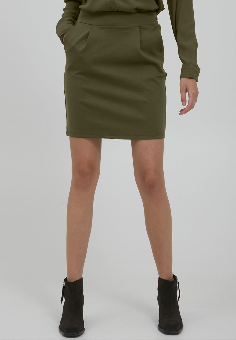 Femme portant une jupe vert olive et un haut assorti avec des bottines noires, debout avec une main dans la poche devant un fond uni.