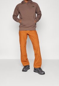Felpa con cappuccio marrone, con tasca frontale e logo, abbinata a pantaloni arancioni e scarpe da esterno grigie con accenti neri.