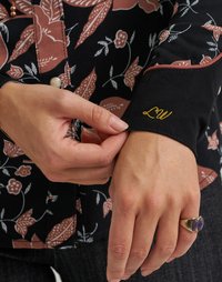 Svart blomstret skjorte med brune bladmønstre. Mansjetten har gull "LW" broderi. Hånd som justerer mansjetten, iført en ring med to steiner.