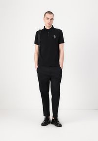 Fekete polo póló, kis hímzett grafikával, elegáns fekete nadrággal és fekete cipővel párosítva. A modell semleges háttér előtt áll.