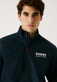 Veste légère navy avec un col montant, fermeture zippée intégrale, et logos imprimés "RBR" et "RED BULL RACING" en blanc sur la poitrine.