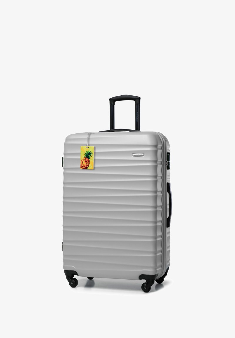 Srebrna twarda walizka z żłobioną powierzchnią, wysuwaną rączką, czarnymi kółkami i kolorową metką do bagażu w kształcie ananasa.
