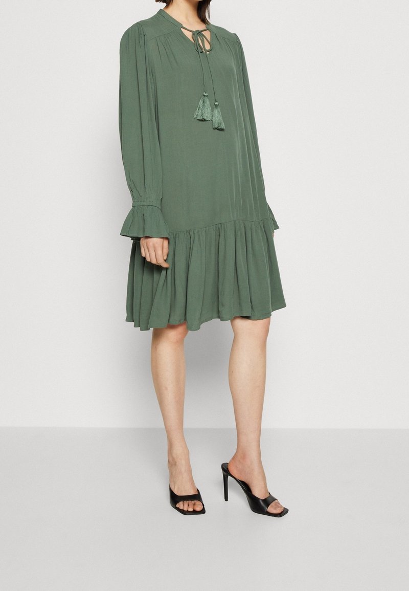 Vestido verde de longitud hasta la rodilla con una tela fluida, mangas acampanadas y un dobladillo en capas; presenta un lazo en el cuello con borlas. Llevado con tacones altos negros.