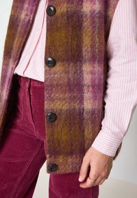 Gilet en laine aux tons terreux, avec un motif à carreaux, six boutons noirs, porté sur une chemise à rayures roses et un pantalon en velours côtelé bordeaux.