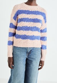 Personne portant un pull en maille rayé rose et bleu associé à un jean denim bleu clair taille haute, debout devant un fond uni.