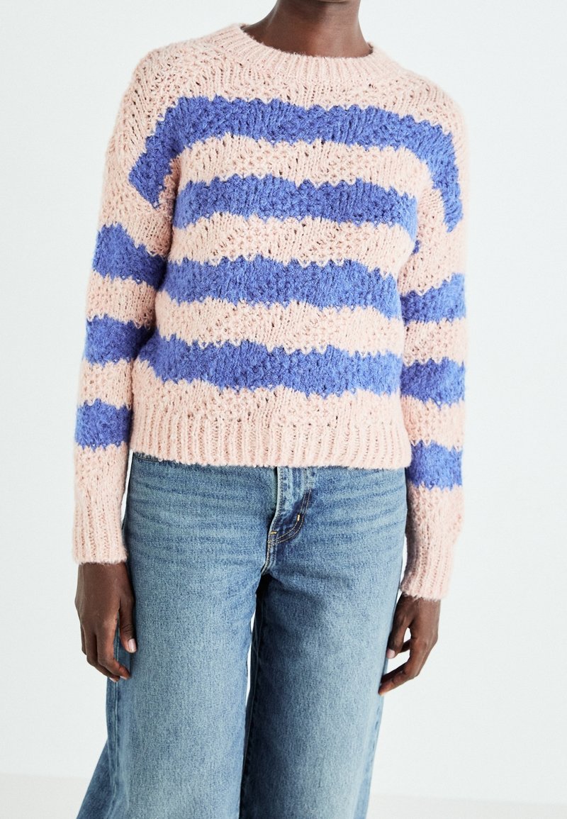Personne portant un pull en maille rayé rose et bleu associé à un jean denim bleu clair taille haute, debout devant un fond uni.