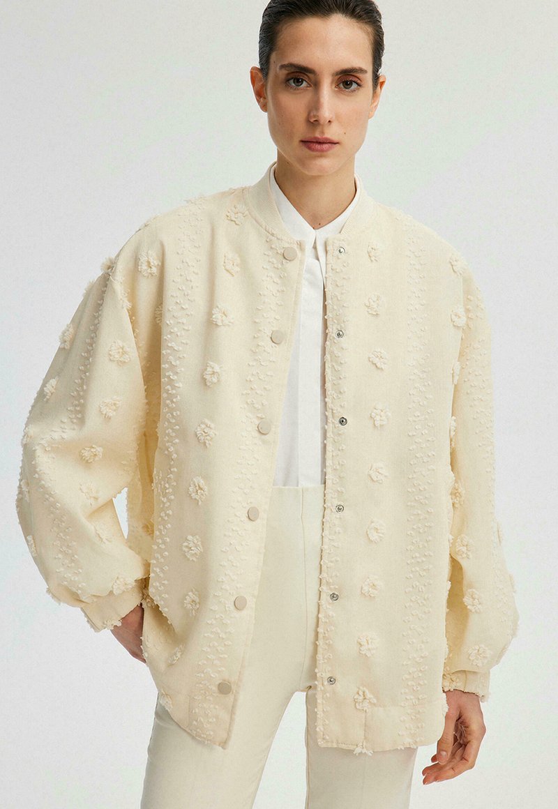 Veste bomber couleur crème avec motifs floraux en relief texturé et fermeture à boutons-pression, présentant une coupe décontractée et un col côtelé.