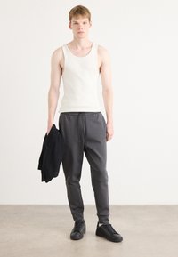 Hvid ribbet tanktop, grå sweatpants med elastik, mørk sweatshirt i hånden og sorte sneakers på et lyst betongulv.