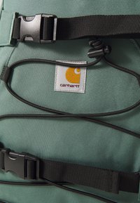 Rucsac din material verde, dotat cu o curea neagră și un sistem de cataramă, având un ecuson cu brand care afișează un logo galben. Design atent elaborat.