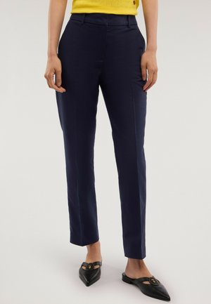 Donna che indossa pantaloni blu scuro su misura, maglia gialla lavorata a maglia e scarpe slingback nere con punta e emblema dorato, in piedi davanti a uno sfondo neutro.