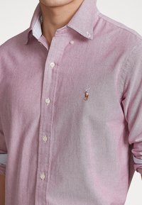 Jasnoróżowa koszula zapinana na guziki, wykonana z miękkiego materiału, z klasycznym kołnierzykiem i białymi oraz jasnoniebieskimi paskami przy mankietach; haftowane logo na piersi.