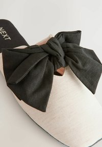 Scarpa slip-on con una toma superiore testurizzata di un beige chiaro, caratterizzata da un grande fiocco in tessuto grigio scuro nella parte anteriore e una soletta nera.