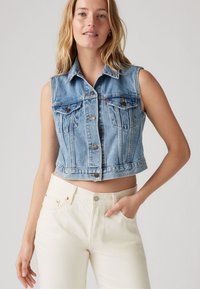 Γυναίκα που φοράει αμάνικο μπλε τζιν γιλέκο με κουμπιά και λευκό ψηλόμεσο τζιν, στέκεται μπροστά σε απλό φωτεινό φόντο.
