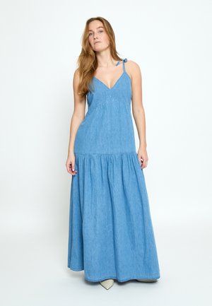 Žena s dlhými vlasmi v modrých dlhých šatách bez rukávov s viazanými ramennými popruhmi, stojaca pred jednoduchým bielym pozadím.