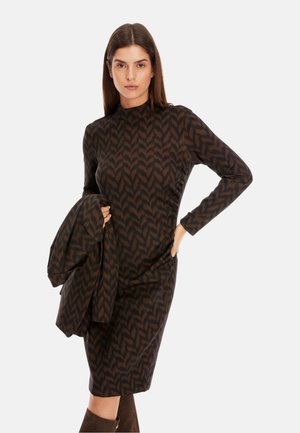 Langärmliges braunes Kleid mit einem schwarzen Zickzackmuster, verfügt über einen hohen Ausschnitt und eine enge Passform. Das Model hält einen passenden Schal.