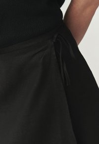 Čierna ľanová sukňa s detailom viazania v páse, s hladkou textúrou a minimalistickým dizajnom. Tkanina jemne padá a elegantne sa drapí.