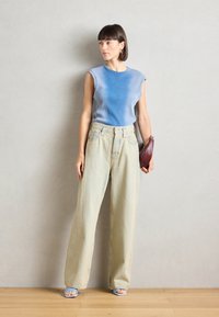 Top sans manches bleu clair à côtes, associé à un jean large vert pâle et ample. Les accessoires comprennent un petit sac à main rouge foncé et des sandales à talons bleues.